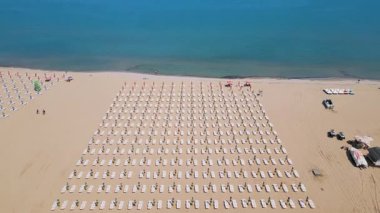 Durgun denize bakan güneş yatakları ve renkli şemsiyeleri olan mükemmel düzenlenmiş bir sahil manzarası. Yukarıdan gelen barışçıl yaz tatili sahnesi, insansız hava aracı tarafından yakalandı..