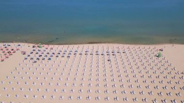 Durgun denize bakan güneş yatakları ve renkli şemsiyeleri olan mükemmel düzenlenmiş bir sahil manzarası. Yukarıdan gelen barışçıl yaz tatili sahnesi, insansız hava aracı tarafından yakalandı..