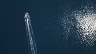Koyu mavi suda hızla ilerleyen bir jet ski 'nin havadan görünüşü, dinamik beyaz bir iz bırakıyor. Yukarıdan insansız hava aracı tarafından ele geçirildi. Denizde hareketlilik, güç ve yaz eğlencesi sergileniyor..