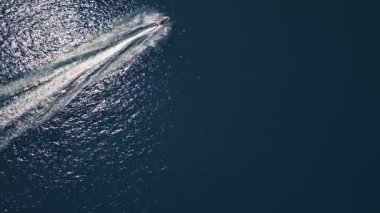 Koyu mavi suda hızla ilerleyen bir jet ski 'nin havadan görünüşü, dinamik beyaz bir iz bırakıyor. Yukarıdan insansız hava aracı tarafından ele geçirildi. Denizde hareketlilik, güç ve yaz eğlencesi sergileniyor..
