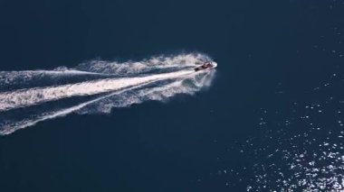 Koyu mavi suda hızla ilerleyen bir jet ski 'nin havadan görünüşü, dinamik beyaz bir iz bırakıyor. Yukarıdan insansız hava aracı tarafından ele geçirildi. Denizde hareketlilik, güç ve yaz eğlencesi sergileniyor..