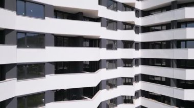 İnsansız hava aracı, beyaz ve koyu gri aksanlı, geometrik balkon ve geniş pencereli modern bir konut binasının ön cephesinde uçuyor..