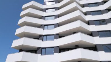 İnsansız hava aracı, keskin açılı balkonları ve açık mavi gökyüzünün altında beyaz minimalist mimarisi olan modern bir apartman cephesi boyunca uçuyor..