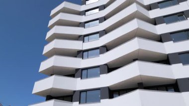 İnsansız hava aracı, keskin açılı balkonları ve açık mavi gökyüzünün altında beyaz minimalist mimarisi olan modern bir apartman cephesi boyunca uçuyor..