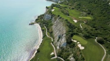 Yeşil çim yolları, kum sığınakları, golf arabaları ve oyuncularla dolu, yemyeşil bitki örtüsü ve turkuaz deniz manzaralı bir sahil golf sahası manzarası..