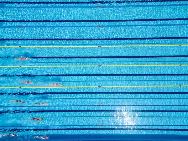 Olimpik boyutta bir havuzda yüzücülerin eğitimini yukarıdan aşağıya gösteren bir görüntü. Hareket halindeki insanlarla dolu bir sürü şerit. Açık mavi suyla aktif yaz sporları ve fitness sahnesi.