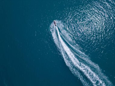 Koyu mavi suda hızla ilerleyen bir jet ski 'nin havadan görünüşü, dinamik beyaz bir iz bırakıyor. Yukarıdan insansız hava aracı tarafından ele geçirildi. Denizde hareketlilik, güç ve yaz eğlencesi sergileniyor..