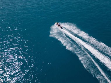 Koyu mavi suda hızla ilerleyen bir jet ski 'nin havadan görünüşü, dinamik beyaz bir iz bırakıyor. Yukarıdan insansız hava aracı tarafından ele geçirildi. Denizde hareketlilik, güç ve yaz eğlencesi sergileniyor..