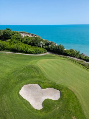 Yeşil çim yolları, kum sığınakları, golf arabaları ve oyuncularla dolu, yemyeşil bitki örtüsü ve turkuaz deniz manzaralı bir sahil golf sahası manzarası..