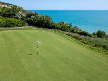 Yeşillik ve sakin turkuaz sularla çevrili, deniz kenarındaki kayalıklarda kareli bir bayrakla golf sahasının yakın görüntüsü..