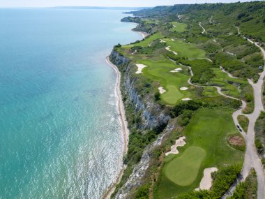 Yeşil çim yolları, kum sığınakları, golf arabaları ve oyuncularla dolu, yemyeşil bitki örtüsü ve turkuaz deniz manzaralı bir sahil golf sahası manzarası..