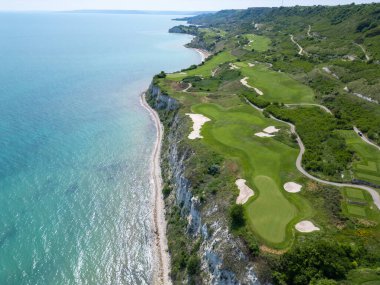 Yeşil çim yolları, kum sığınakları, golf arabaları ve oyuncularla dolu, yemyeşil bitki örtüsü ve turkuaz deniz manzaralı bir sahil golf sahası manzarası..
