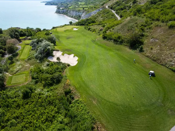 Yeşil çim yolları, kum sığınakları, golf arabaları ve oyuncularla dolu, yemyeşil bitki örtüsü ve turkuaz deniz manzaralı bir sahil golf sahası manzarası..