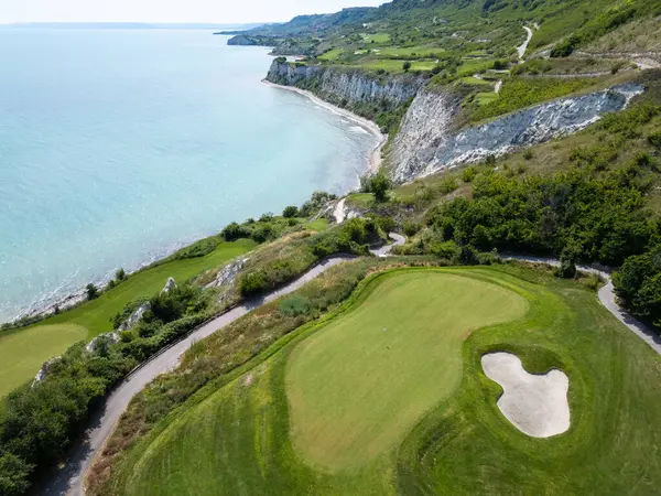 Yeşil çim yolları, kum sığınakları, golf arabaları ve oyuncularla dolu, yemyeşil bitki örtüsü ve turkuaz deniz manzaralı bir sahil golf sahası manzarası..