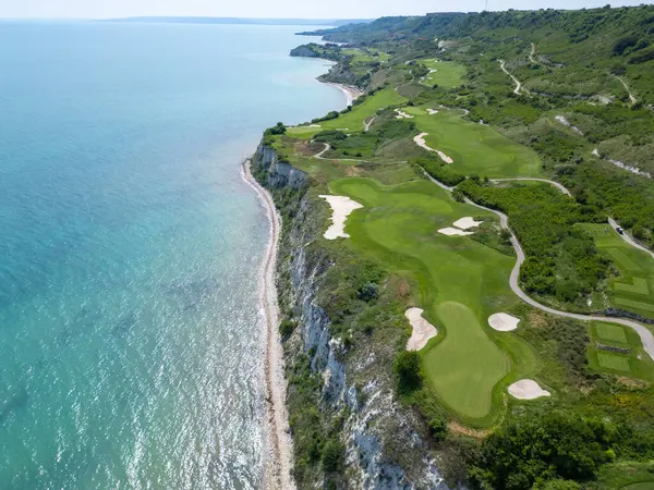 Yeşil çim yolları, kum sığınakları, golf arabaları ve oyuncularla dolu, yemyeşil bitki örtüsü ve turkuaz deniz manzaralı bir sahil golf sahası manzarası..