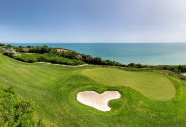Yeşil çim yolları, kum sığınakları, golf arabaları ve oyuncularla dolu, yemyeşil bitki örtüsü ve turkuaz deniz manzaralı bir sahil golf sahası manzarası..