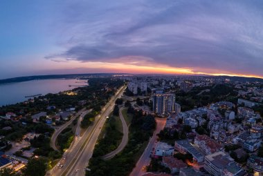 Bulgaristan 'ın Varna kentinde günbatımında havadan panoramik manzara. Karadeniz üzerinde kıyı kenti ışıkları, konut binaları, otoyollar ve dramatik gökyüzü bulunan şehir manzarası.