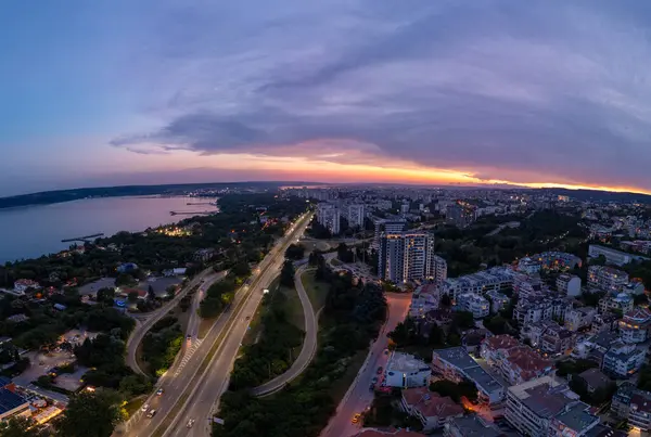Bulgaristan 'ın Varna kentinde günbatımında havadan panoramik manzara. Karadeniz üzerinde kıyı kenti ışıkları, konut binaları, otoyollar ve dramatik gökyüzü bulunan şehir manzarası.