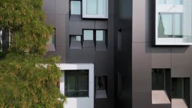 Geometrik siyah panelleri ve çıkıntılı pencereleri olan modern bir binanın yakın çekim videosu. Yeşil bir ağaç kontrastı ekler. Temiz çizgiler ve cam elementlerle minimalist mimari.