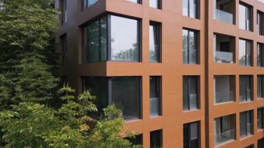 Çevresi yeşil ağaçlarla çevrili, geniş pencereli ve kahverengi cepheli modern apartman binası. Barışçıl şehir ortamında çağdaş mimari. Gündüz görüşü, insan yok..