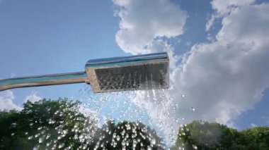 Açık havada duş alan su damlacıkları parlak güneşin altında ağaçlar ve arkadaki mavi gökyüzü ile taze bir yaz anını yakalıyor..