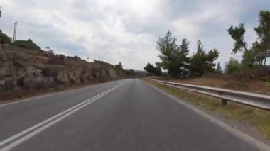 Hareket halindeki bir arabadan manzaralı kıyı yolu manzarası, bir tarafında deniz, bir tarafında kayalık uçurumlar, diğer tarafında kuru çimenler ve ağaçlar parçalı bulutlu bir gökyüzünün altında.