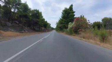 Hareket halindeki bir arabadan manzaralı kıyı yolu manzarası, bir tarafında deniz, bir tarafında kayalık uçurumlar, diğer tarafında kuru çimenler ve ağaçlar parçalı bulutlu bir gökyüzünün altında.