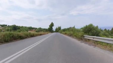 Hareket halindeki bir arabadan manzaralı kıyı yolu manzarası, bir tarafında deniz, bir tarafında kayalık uçurumlar, diğer tarafında kuru çimenler ve ağaçlar parçalı bulutlu bir gökyüzünün altında.