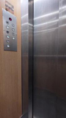 Konut katında asansör kapıları açılıyor, düğmeli ve ışıklı bir zemin numarası olan kontrol panelini gösteriyor. Ilık ışıklandırma ve ahşap kaplama tipik bir iç mekan atmosferi yaratır.