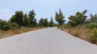 Hareket halindeki bir arabadan manzaralı kıyı yolu manzarası, bir tarafında deniz, bir tarafında kayalık uçurumlar, diğer tarafında kuru çimenler ve ağaçlar parçalı bulutlu bir gökyüzünün altında.