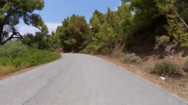 Hareket halindeki bir arabadan manzaralı kıyı yolu manzarası, bir tarafında deniz, bir tarafında kayalık uçurumlar, diğer tarafında kuru çimenler ve ağaçlar parçalı bulutlu bir gökyüzünün altında.