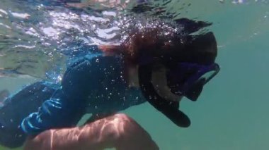 Açık turkuaz suda şnorkelle yüzen, sarı gömlek giyen ve dalış maskesi takan bir çocuğun sualtı selfie 'si. Güneş ışığı ve baloncuklar sahneye eğlenceli ve maceracı bir hava katar..