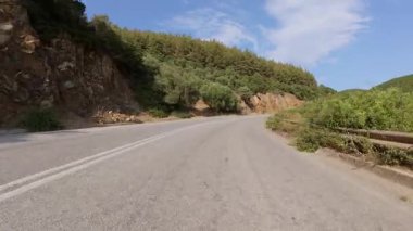 Hareket halindeki bir arabadan manzaralı kıyı yolu manzarası, bir tarafında deniz, bir tarafında kayalık uçurumlar, diğer tarafında kuru çimenler ve ağaçlar parçalı bulutlu bir gökyüzünün altında.