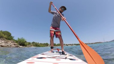 Açık mavi gökyüzünün altındaki yeşil kıyı şeridi yakınındaki temiz sığ suda kürek çeken bir adam. Renkli raket ve pano ile aktif yaz yaşam tarzı sahnesi, açık hava macera temaları için mükemmel.