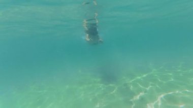 Çocuk sığ ve berrak denizde yüzüyor. Bir şnorkel maskesi ve yüzme şortu takarak, baloncuklar ve güneş ışığı desenleriyle çevrili kumlu okyanus tabanını keşfeder..