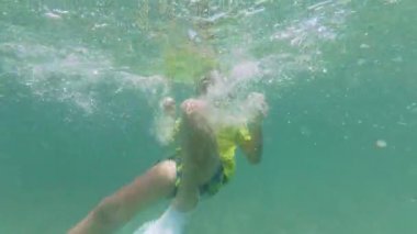 Çocuk sığ ve berrak denizde yüzüyor. Bir şnorkel maskesi ve yüzme şortu takarak, baloncuklar ve güneş ışığı desenleriyle çevrili kumlu okyanus tabanını keşfeder..