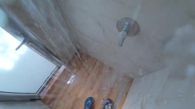 Bir insanın duş alırken, mavi sandaletlere su sıçrarken, fayanslı bir banyoda ahşap bir zeminde, gün ışığı buzlu bir pencereden içeri girerken görüntüsü..