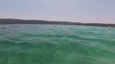 Uzak ormanlık tepeleri ve ufukta bir yatı olan turkuaz deniz suyunun manzarası. Sakin dalgalar ve parlak yaz gökyüzü huzurlu bir tatil ortamı yaratır..