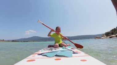 Gülümseyen bir çocuk, turkuaz deniz üzerinde ayakta duran bir kürek çekiyor. Doğada yaz macerası, sörf tahtasında paletler ve arka planda manzaralı kıyı şeridi..