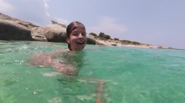Güneşli bir yaz gününde Rocky Beach yakınlarında kristal berraklığında yüzen gülümseyen çocuk. Mutlu çocuk doğada tatilin tadını çıkarıyor, yarı dalmış kameraya bakıyor..