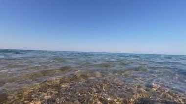 Yumuşak deniz dalgalarının açık sualtı çakıl taşlarını ve mavi gökyüzünü gösteren ve kıyı şeridinin sakin güzelliğini yakalayan bölünmüş seviyedeki videosu..
