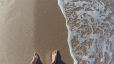 İki çocuğuyla birlikte deniz kıyısında yürüyen bir babanın birinci şahıs görüntüsü. Güneşli bir sahil gününde yumuşak dalgalar gibi çıplak ayaklar ıslak kumda süzülür..