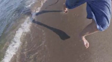 Kumlu bir sahilde, sığ sularda çıplak ayakla koşan bir çocuğun yavaş çekimde güneş ışığı altında dalgaların arasından süzülmesi..