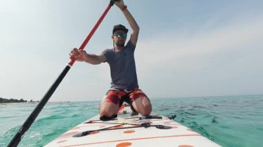 Güneşli bir günde, ayakta kürek çeken bir adam diz çöküp durgun turkuaz suyun tadını çıkarıyor. POV aksiyon kamerası temiz gökyüzü ve ufukta.