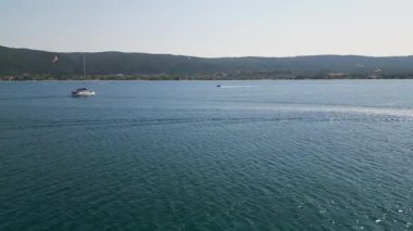 Yelkenli açık gökyüzünün altındaki ormanlık bir sahil şeridinde durgun mavi denizde seyrediyor. Tekneler ve uzak sahil köyüyle huzurlu bir yaz günü..