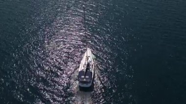 Bir yelkenlinin parlak güneş ışığı altında derin mavi denizde yüzüşünün üst görüntüsü. Suyun parlak yansımaları huzurlu, manzaralı bir deniz atmosferi yaratır..
