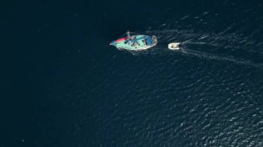 Koyu mavi denizde küçük bir tekneyi çeken renkli bir yelkenlinin hava videosu. Yukarıdan aşağıya deniz hareketlerini, seyahatleri ve deniz yaşamını gösteriyor.