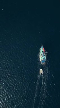 Koyu mavi denizde küçük bir tekneyi çeken renkli bir yelkenlinin hava videosu. Yukarıdan aşağıya deniz hareketlerini, seyahatleri ve deniz yaşamını gösteriyor.