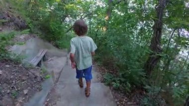 Dar bir orman yolunda yeşil ağaçlar ve yaz bitkileriyle çevrili beton adımlarla yürüyen bir çocuğu izleyen birinci şahıs POV. Ormanda barışçıl doğa keşfi.