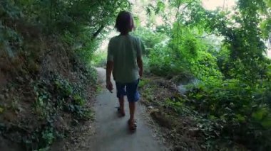 Dar bir orman yolunda yeşil ağaçlar ve yaz bitkileriyle çevrili beton adımlarla yürüyen bir çocuğu izleyen birinci şahıs POV. Ormanda barışçıl doğa keşfi.